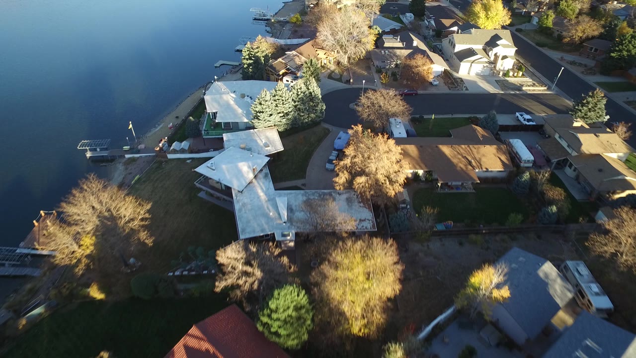Aerial view in Westminster, Colorado with blue lake water on the left, clustered houses and driveways on the right, bare trees, yellow foliage, and no visible people.