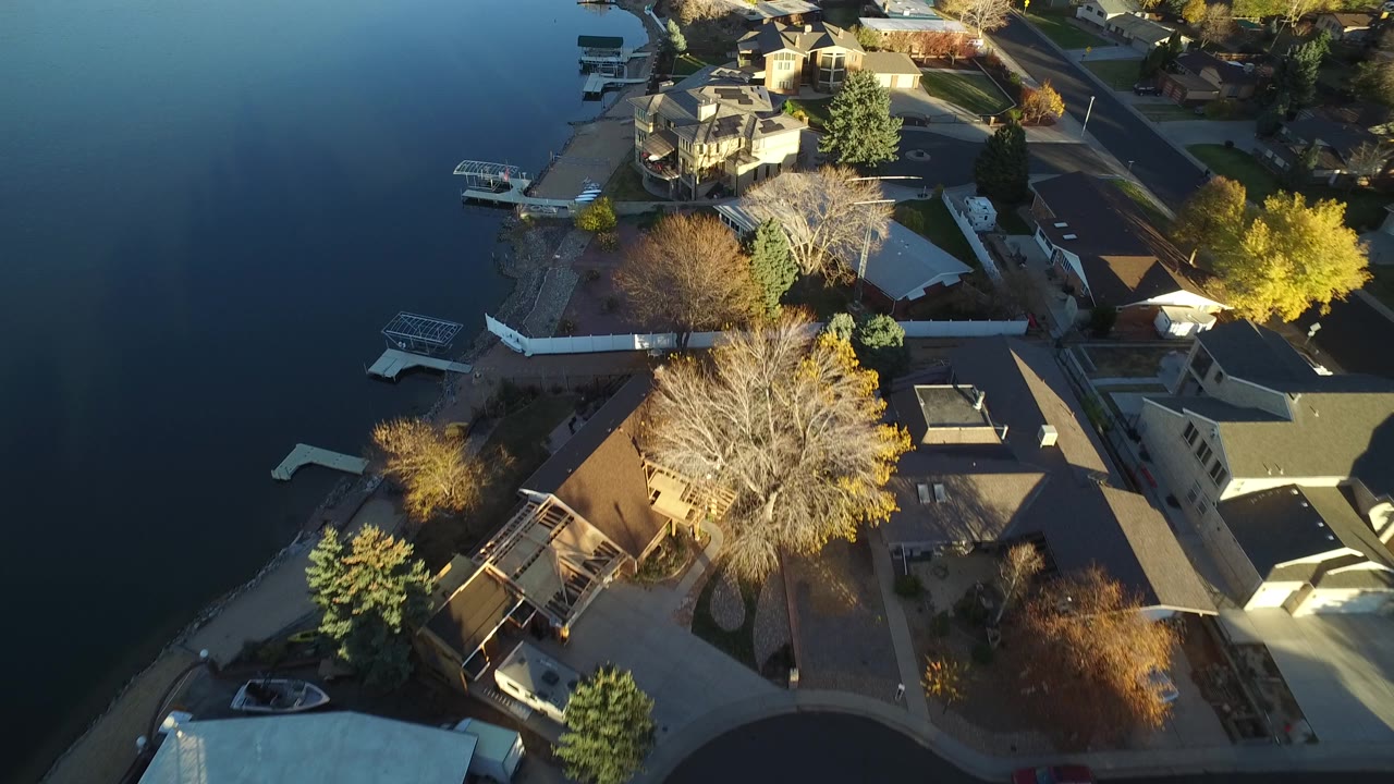 The Westminster aerial continues over dark lake water and shoreline homes, with docks, large trees, rooftops, streets, and no visible people.