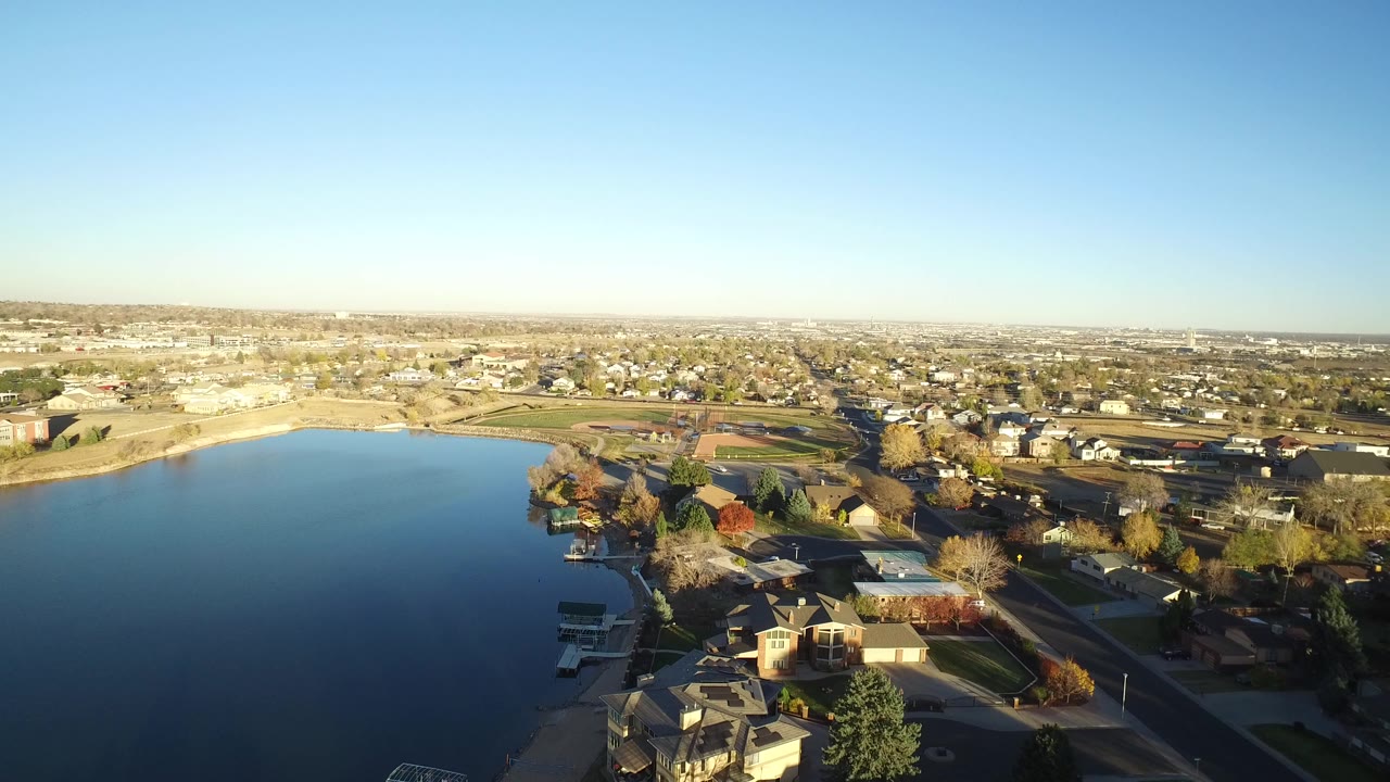 Opening aerial frame over a Westminster, Colorado lake with shoreline homes, autumn-colored trees, open fields, distant suburbs, blue sky, and no visible people.
