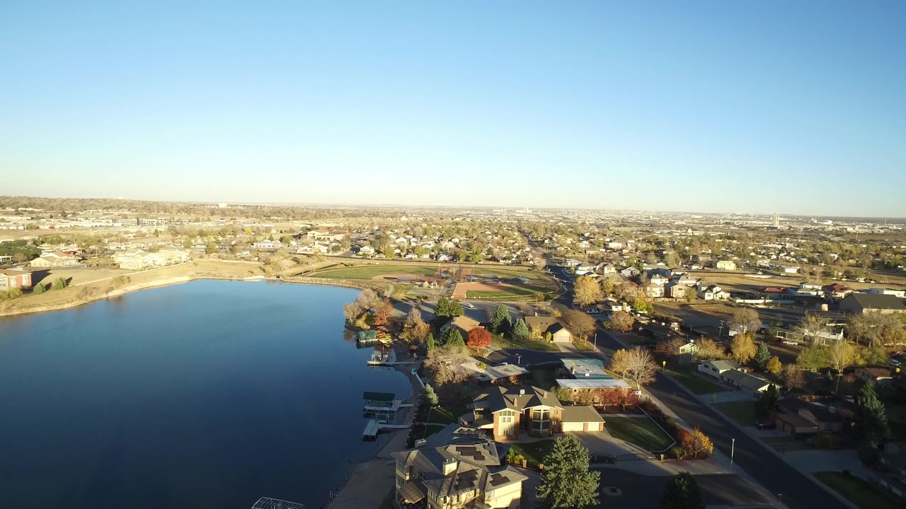 Aerial frame of the Westminster, Colorado lakeshore with dark blue water at left, houses and small docks below, sunlit fields and suburban blocks beyond, and no visible people.