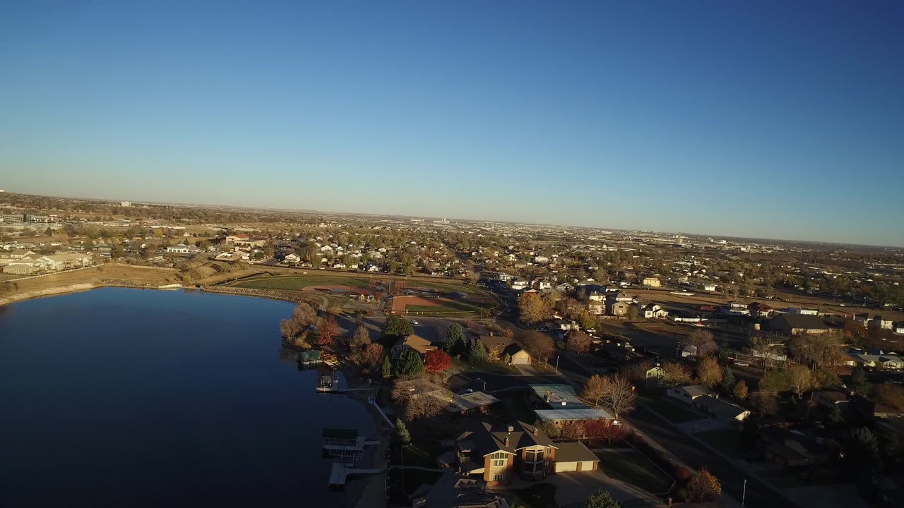 Wide aerial frame over Westminster, Colorado with a dark lake on the left, private homes in the foreground, sports fields beyond, distant city buildings on the horizon, and no visible people.