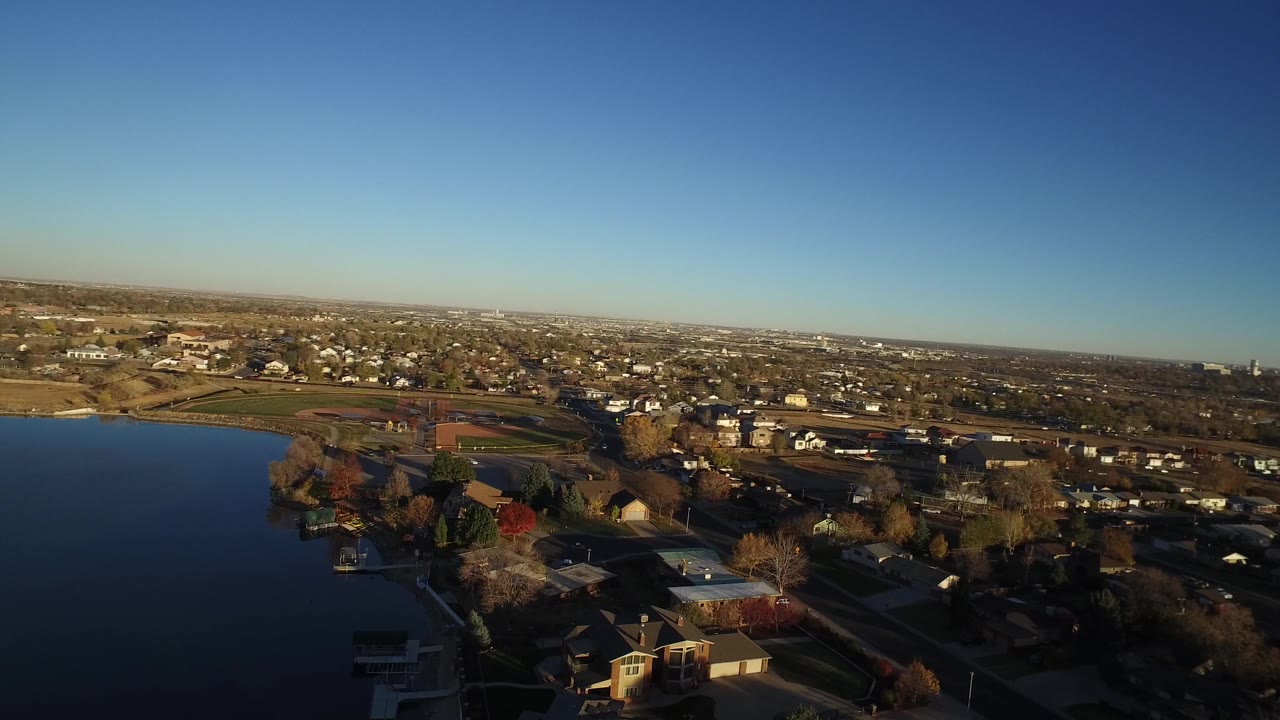 Aerial suburban landscape with lake edge, houses, sports fields, roads, and skyline in the distance.