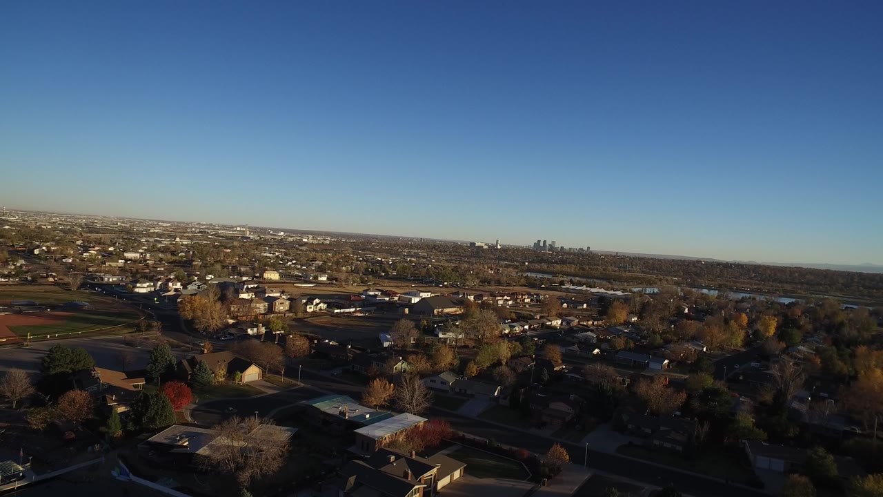 Aerial view over suburban homes, streets, trees, and a distant skyline under blue sky.