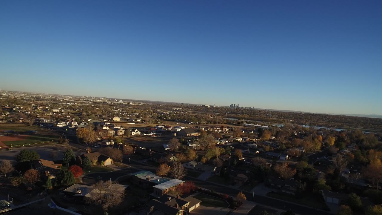 A slightly later aerial view of Westminster, Colorado showing residential blocks, scattered trees, open green areas, distant water, skyline, and no visible people.