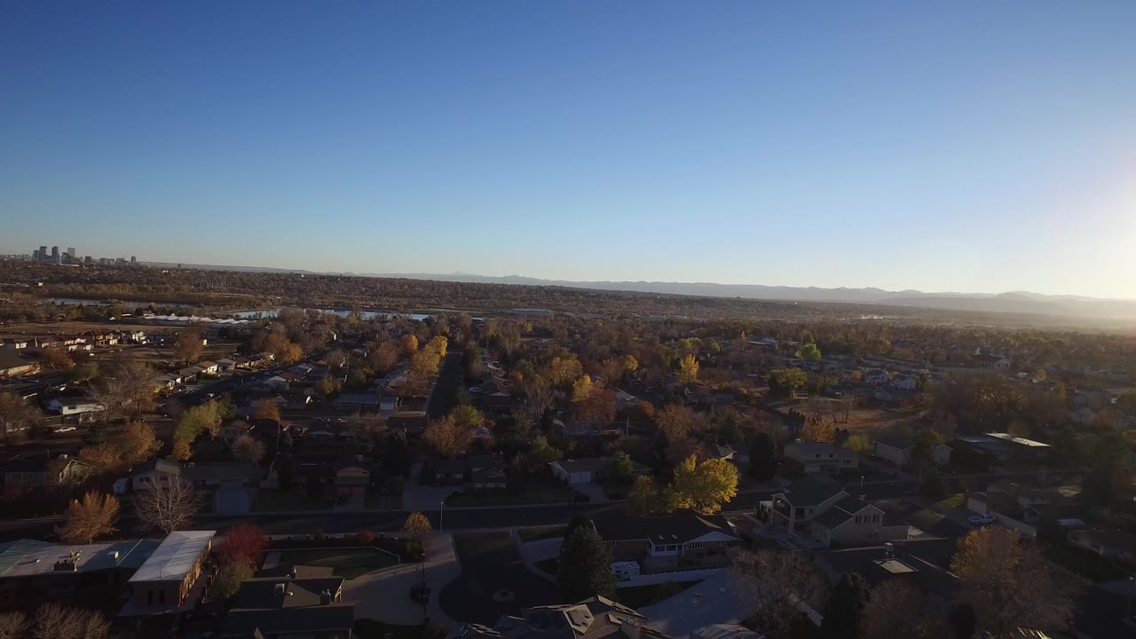 Aerial view over suburban homes and trees with distant skyline and low sun.