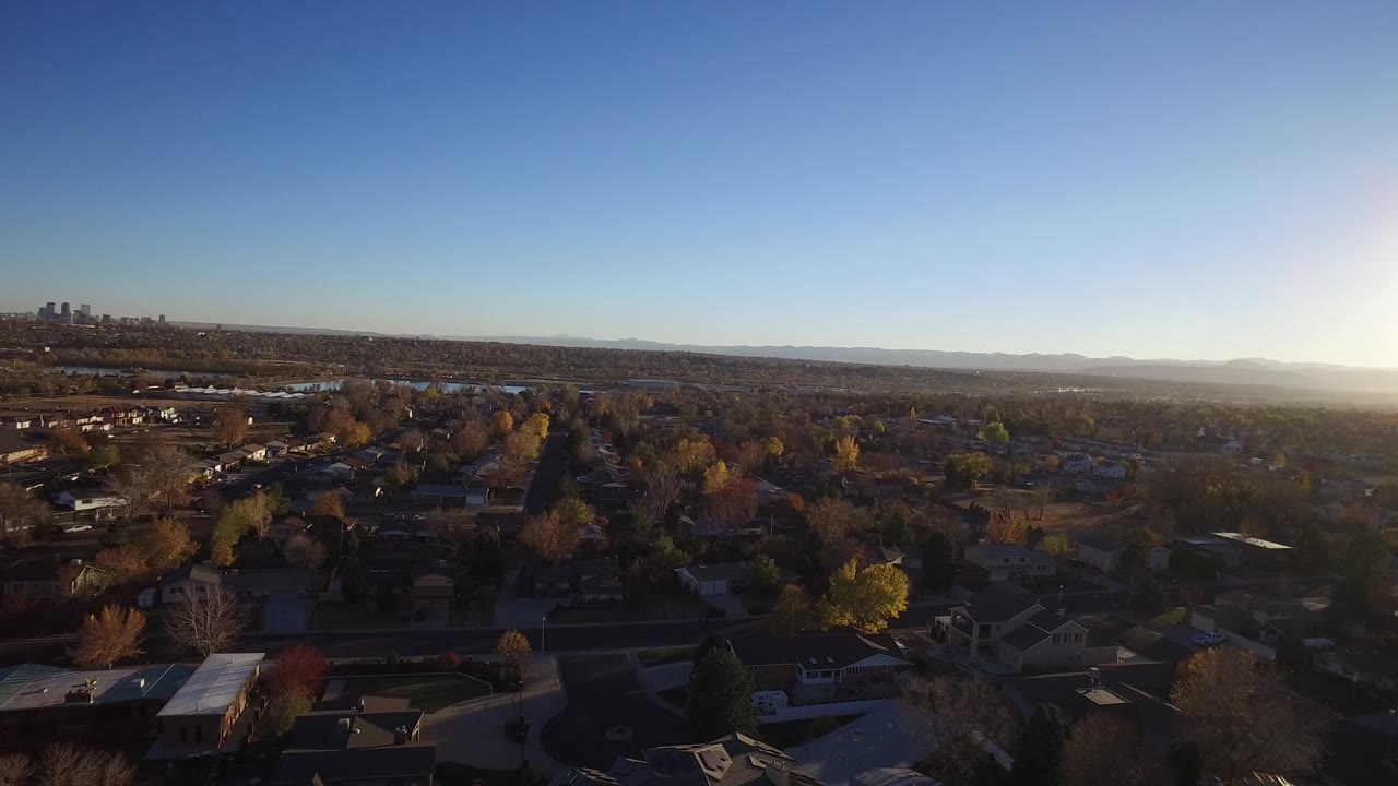 A second aerial frame over Westminster, Colorado shows rooftops and tree-lined blocks under stronger sun flare, with the distant skyline still visible at upper left and no visible people.
