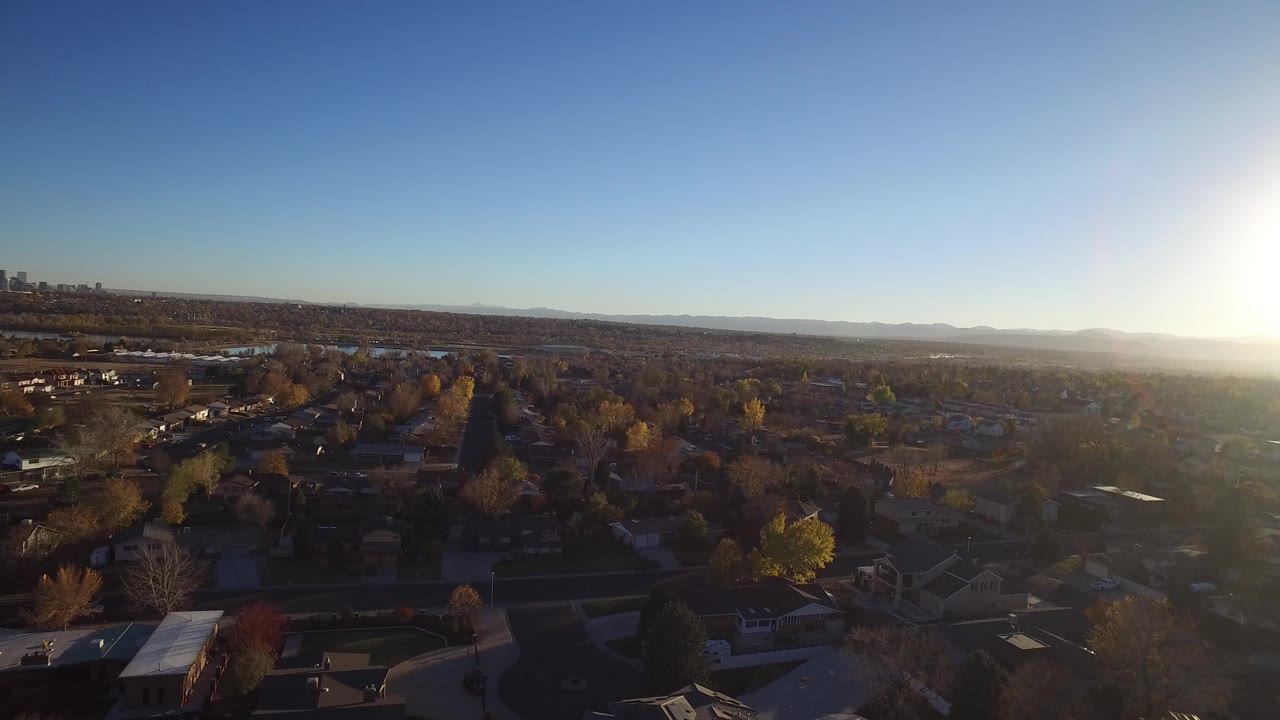 Suburban aerial facing the low sun with homes, trees, and distant mountains.