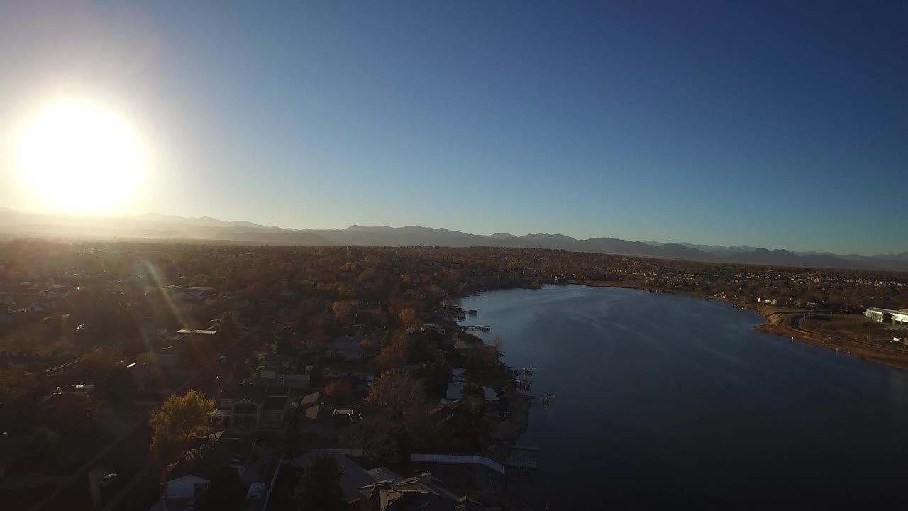 Westminster, Colorado aerial frame with the lake centered, residential rooftops in shadow, distant ridgelines, blue sky, and bright sun glare on the left with no visible people.