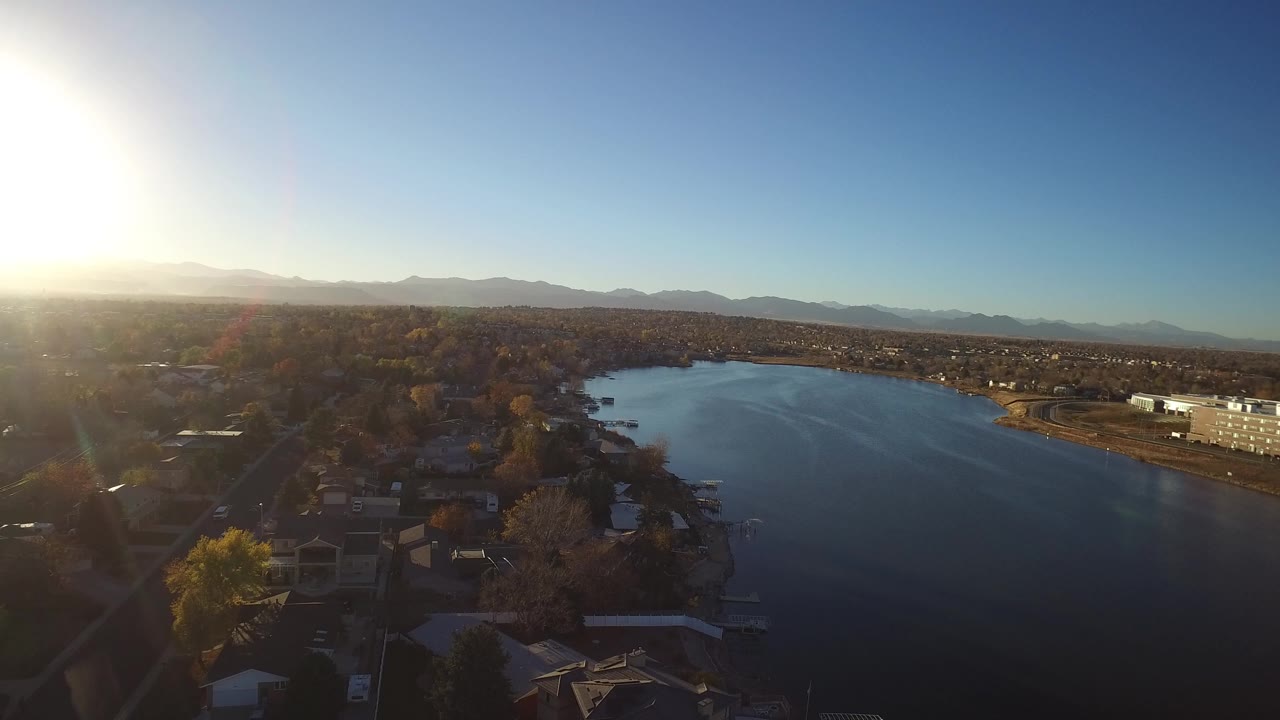 Aerial view of a lake with homes, trees, and distant mountains under bright sun.