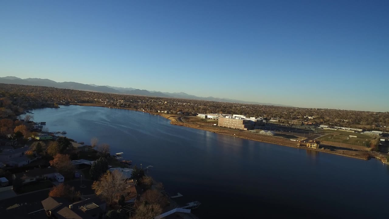 Aerial view in Westminster, Colorado, with the lake filling the lower frame, tree-lined houses along the near shore, pale buildings beyond the water, distant mountains, and no visible people.