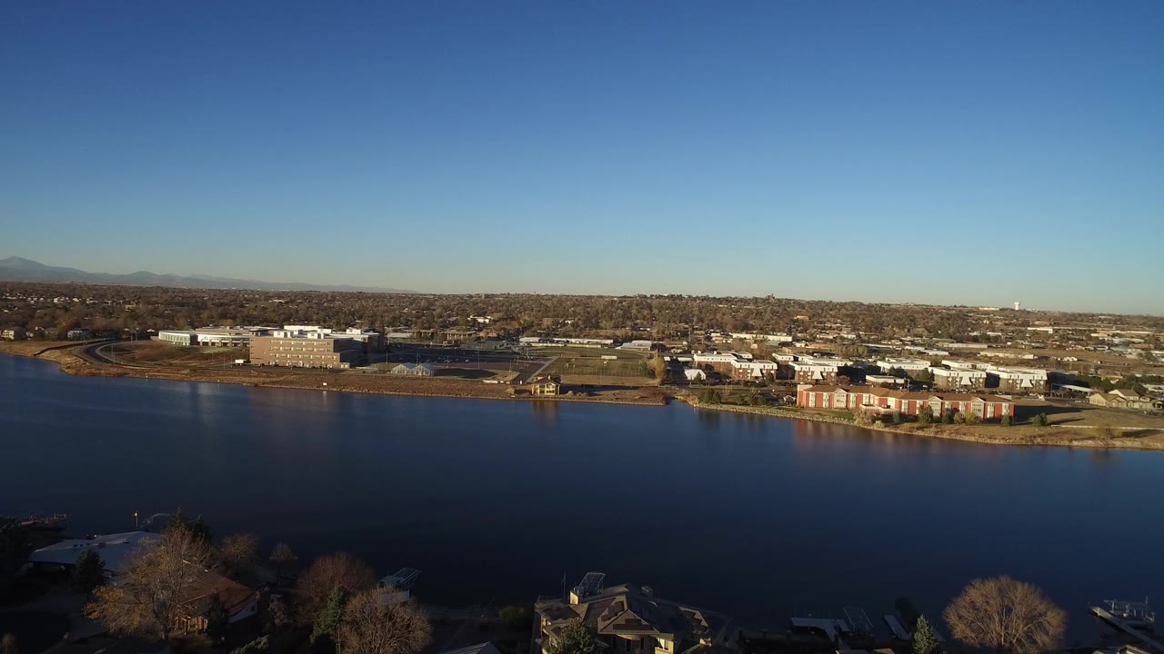 Opening aerial frame in Westminster, Colorado showing blue water in the foreground, a far shoreline with low buildings, distant mountains, clear sky, and no visible people.