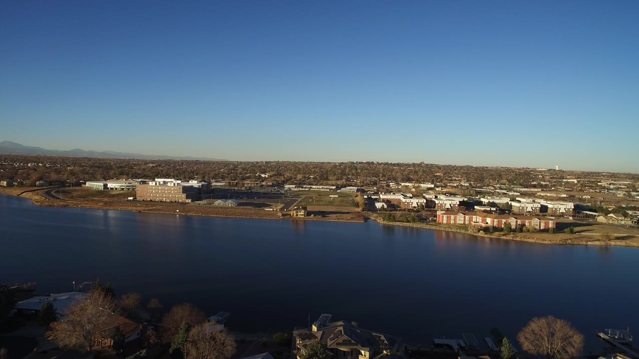 Westminster, Colorado aerial frame with the water surface covering the lower half, leafless foreground trees, far-bank buildings, distant neighborhoods, mountains, and no visible people.