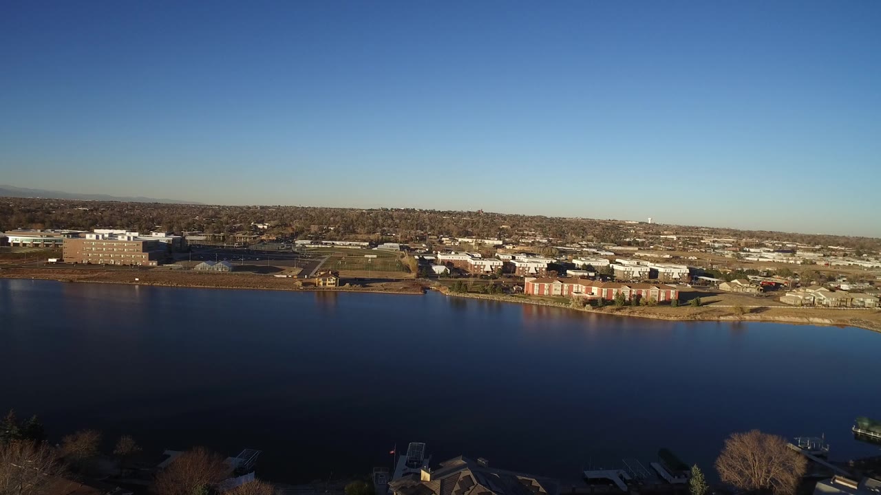 Aerial frame from Westminster, Colorado with broad water in front, a row of shoreline residences and larger low buildings beyond, and no visible people.
