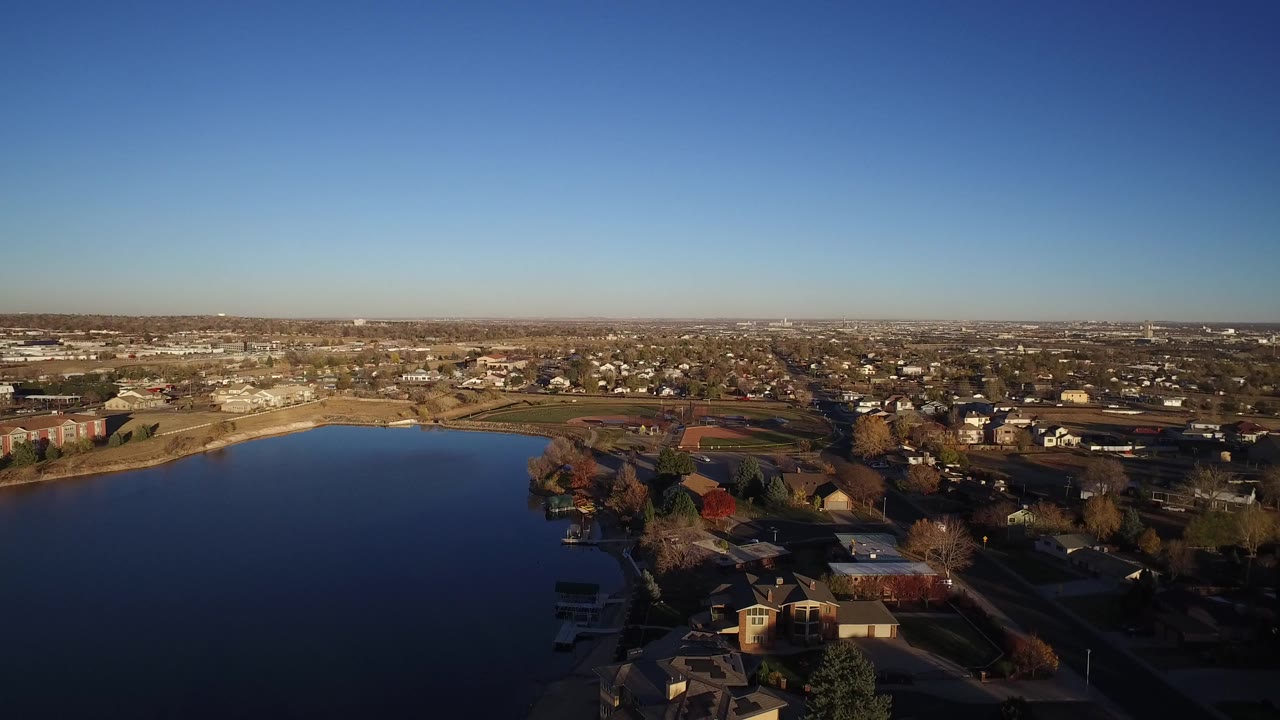 Opening aerial view over Westminster, Colorado with a dark blue lake in the foreground, waterfront homes at lower right, low-rise buildings in the distance, and no visible people.