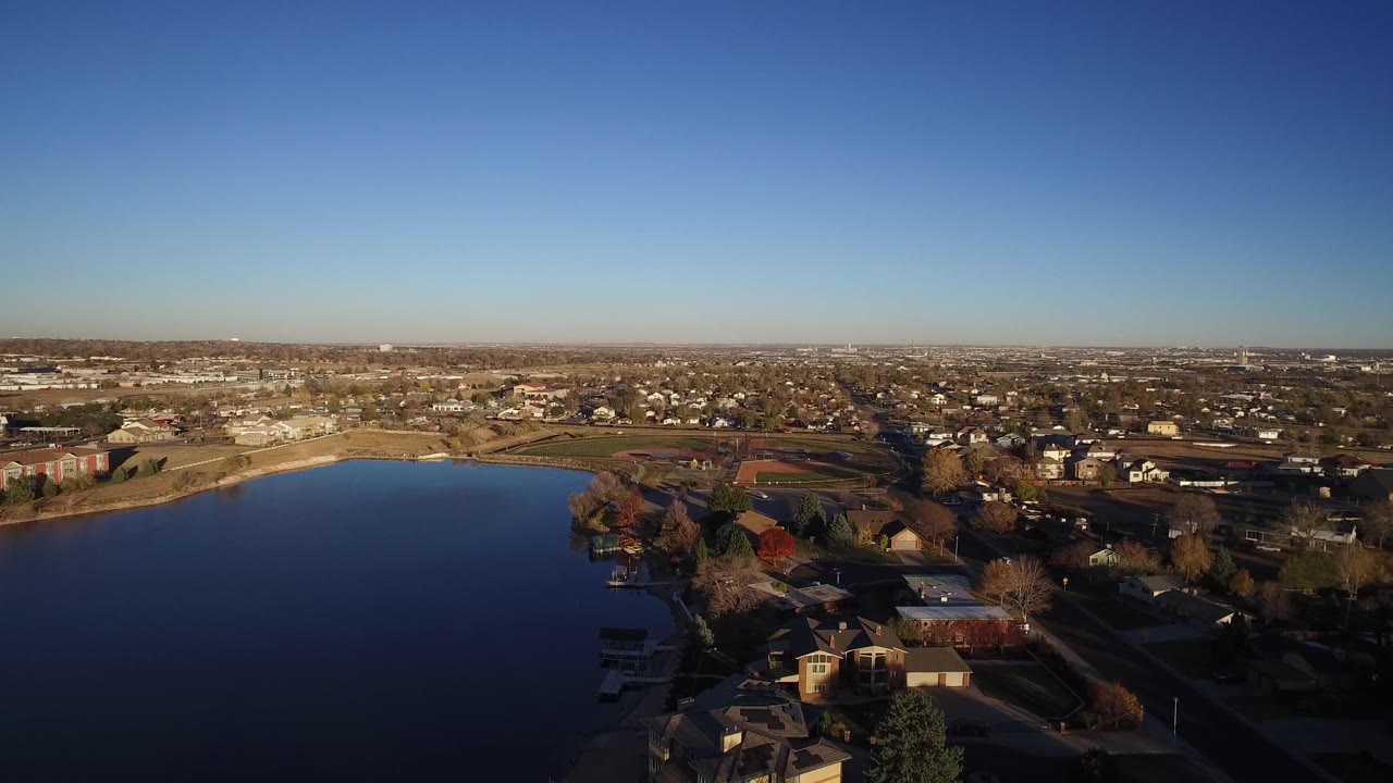 Drone-height view of lakefront houses and a sprawling suburb in bright sunlight.