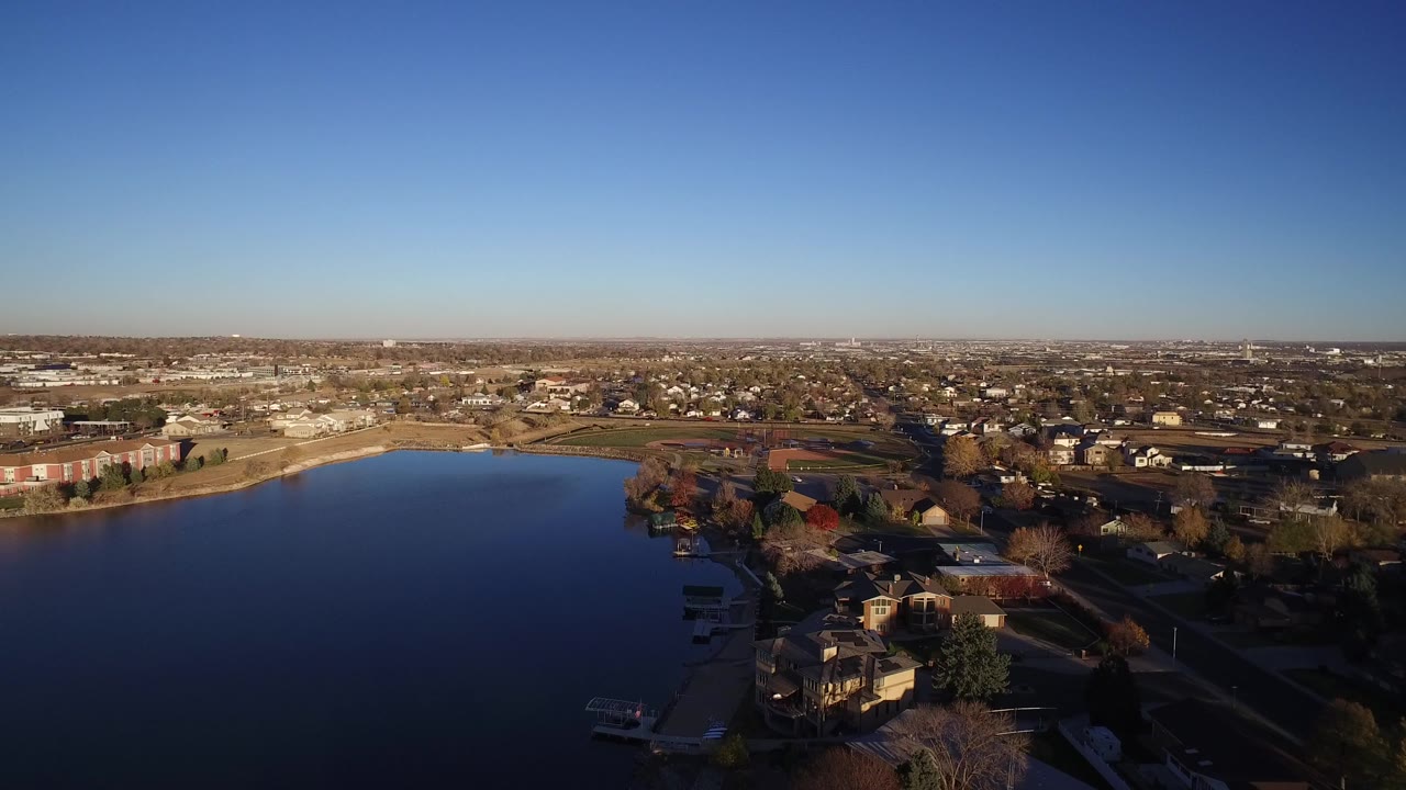 Wide aerial view of a blue lake beside houses and low buildings.