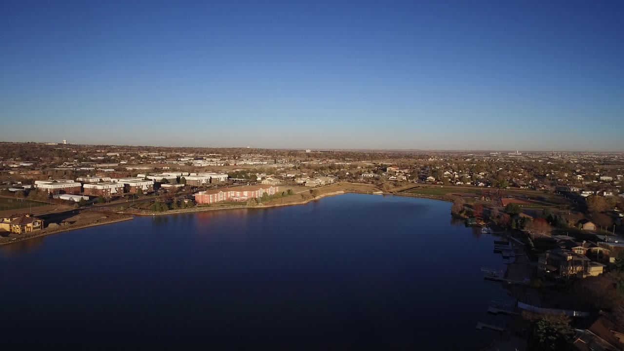 Wide aerial view over a lake toward suburban buildings under a clear blue sky