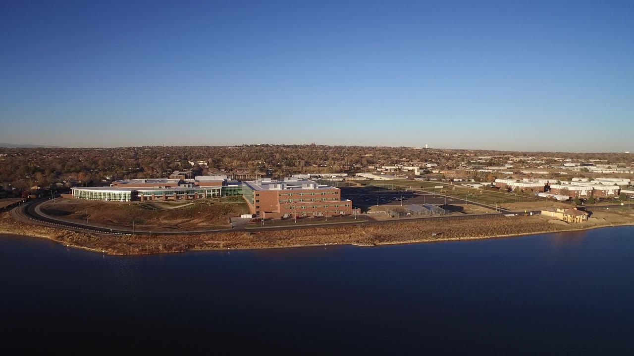 Westminster, Colorado aerial view with the brick building nearer the center, white institutional buildings to the left, a curved shoreline road, open water below, and no visible people.