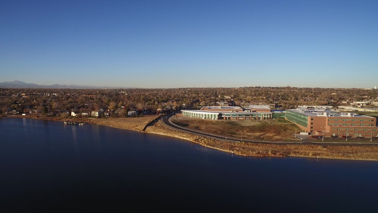Aerial frame in Westminster, Colorado looking across dark water to a curved shoreline, a large low building, nearby tracks or road, suburban houses, distant mountains, and no visible people close enough to identify.
