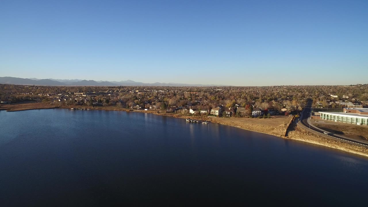 Wide aerial view over dark water toward a residential shoreline and distant mountains.