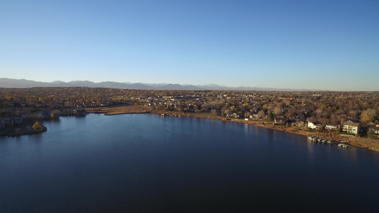 Aerial view over dark lake water toward a suburban shoreline and distant mountains.
