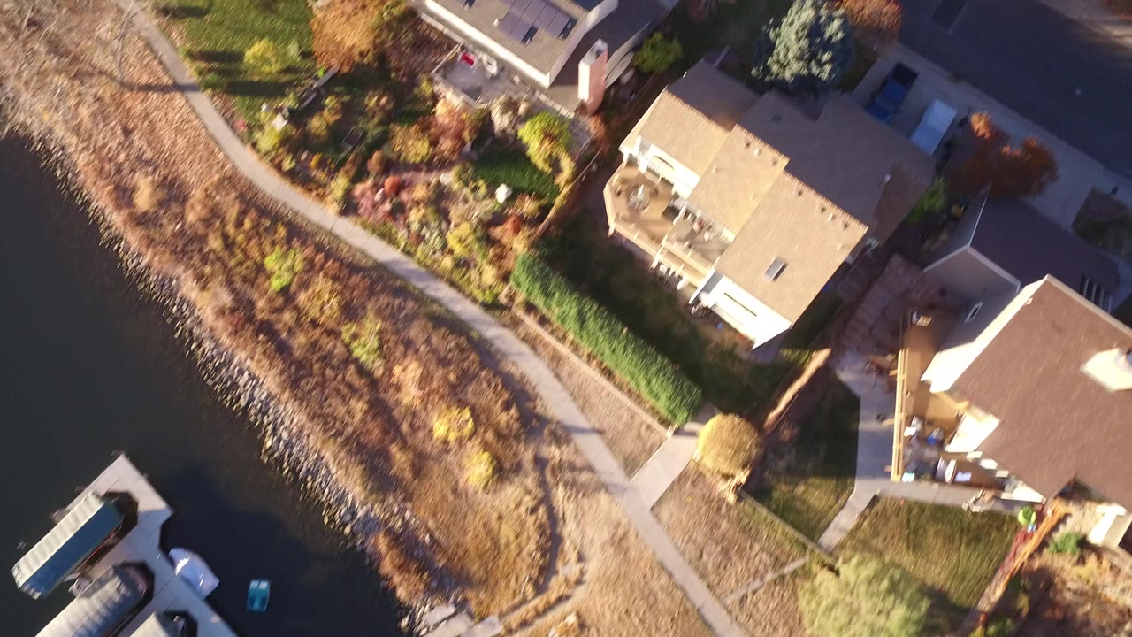 Aerial view in Westminster, Colorado showing detached houses, green lawns, a sidewalk along brown shoreline plants, parked items near a dock, and no visible people.