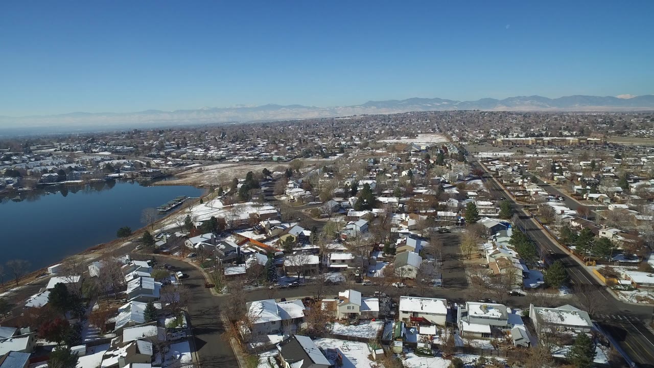 Opening aerial frame over Hidden Lake, Colorado, showing snow-dusted houses, a dark lake on the left, distant mountains, and no visible people.