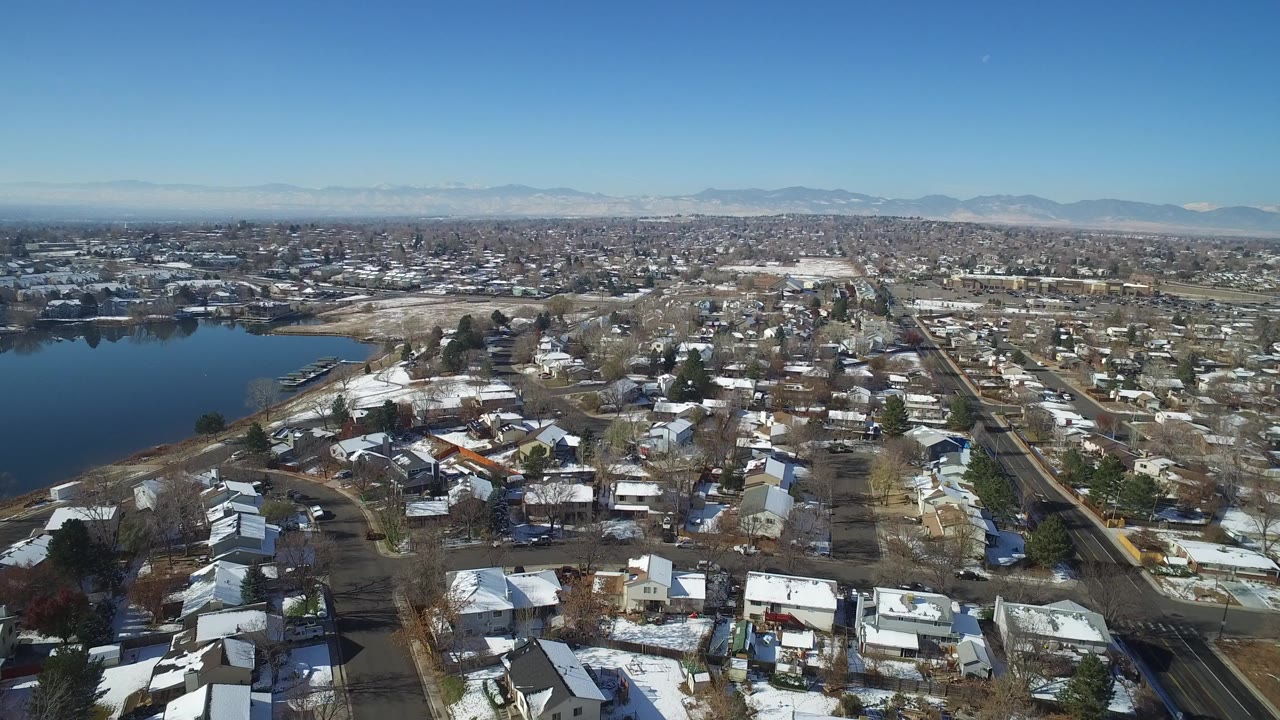 Aerial view of snow-dusted houses, streets, a lake, and distant mountains.