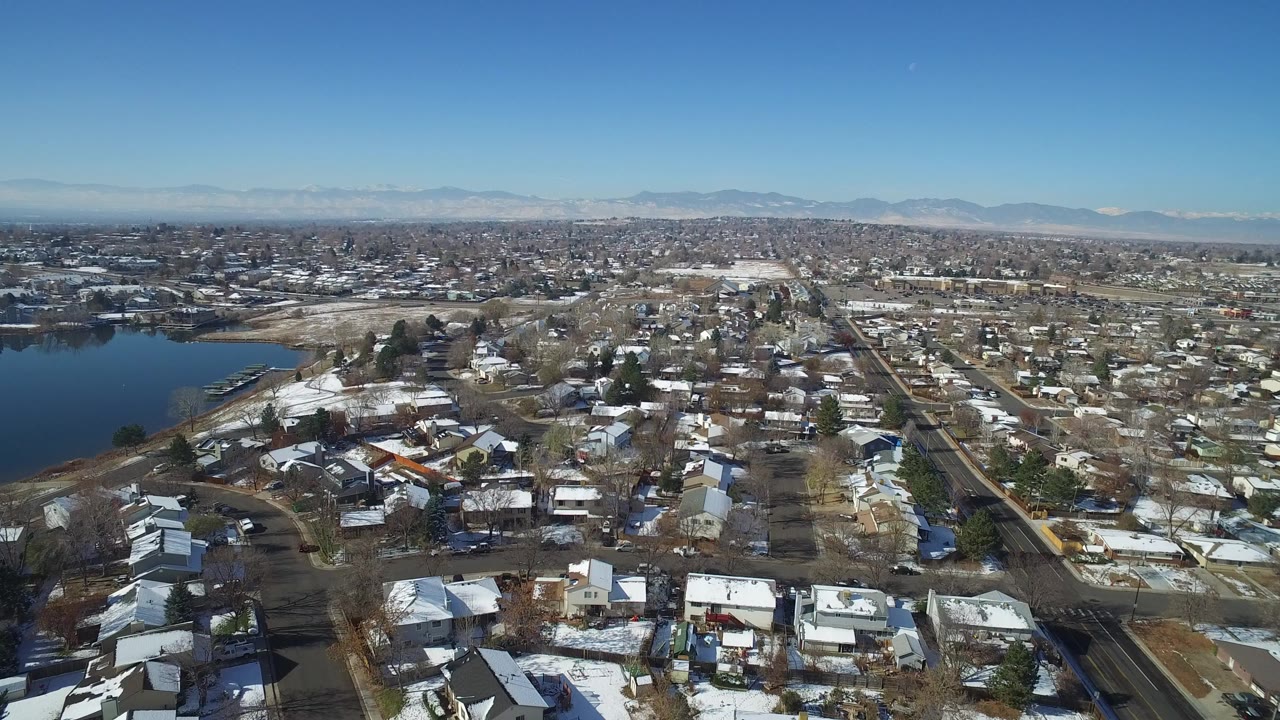 High aerial view over Hidden Lake, Colorado, with snow-dusted homes in the foreground, a dark lake on the left, distant mountains on the horizon, and no visible people.