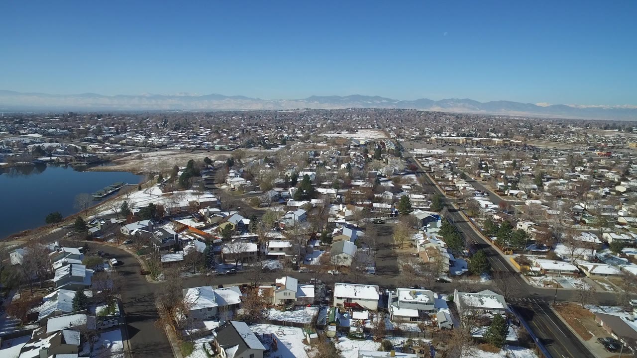 Aerial view over snow-dusted homes, roads, a lake edge, and distant mountains.