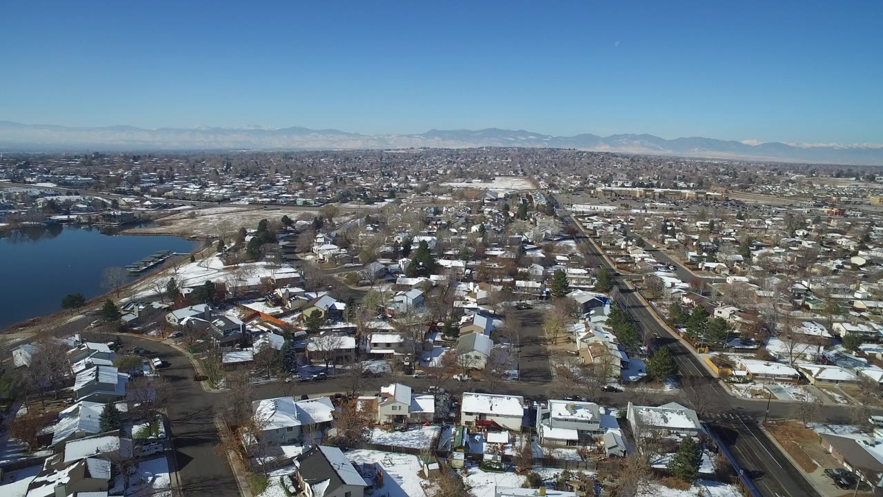 Opening aerial frame over Hidden Lake, Colorado homes with patchy snow, a lake along the left edge, distant mountains on the horizon, and no visible people.