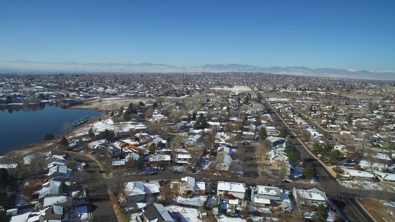High-angle Hidden Lake, Colorado view showing rooftops in the foreground, the lake to the left, long streets through the neighborhood, mountain silhouettes, and no visible people.