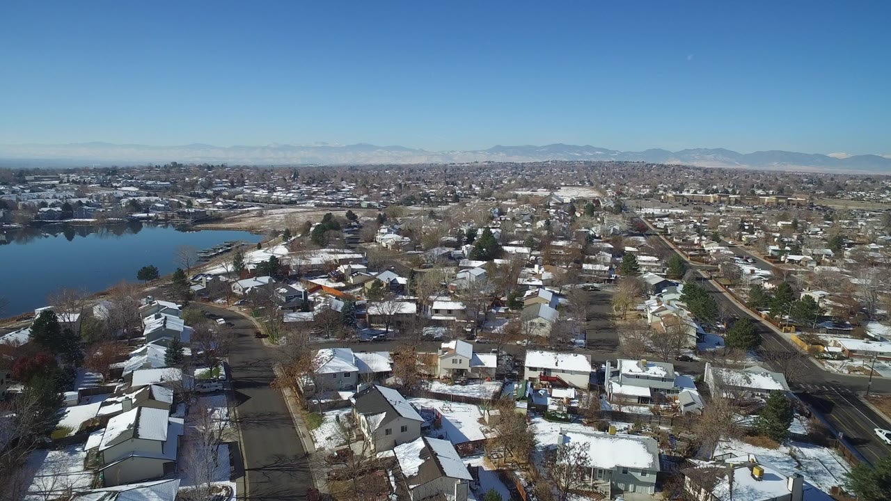 Aerial view of snowy houses, streets, pond, and distant mountains.