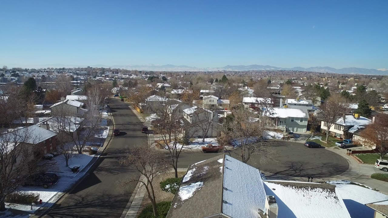 Opening aerial frame over Hidden Lake, Colorado, with a foreground roof, snow patches on lawns, bare trees, distant mountains, and no visible people.