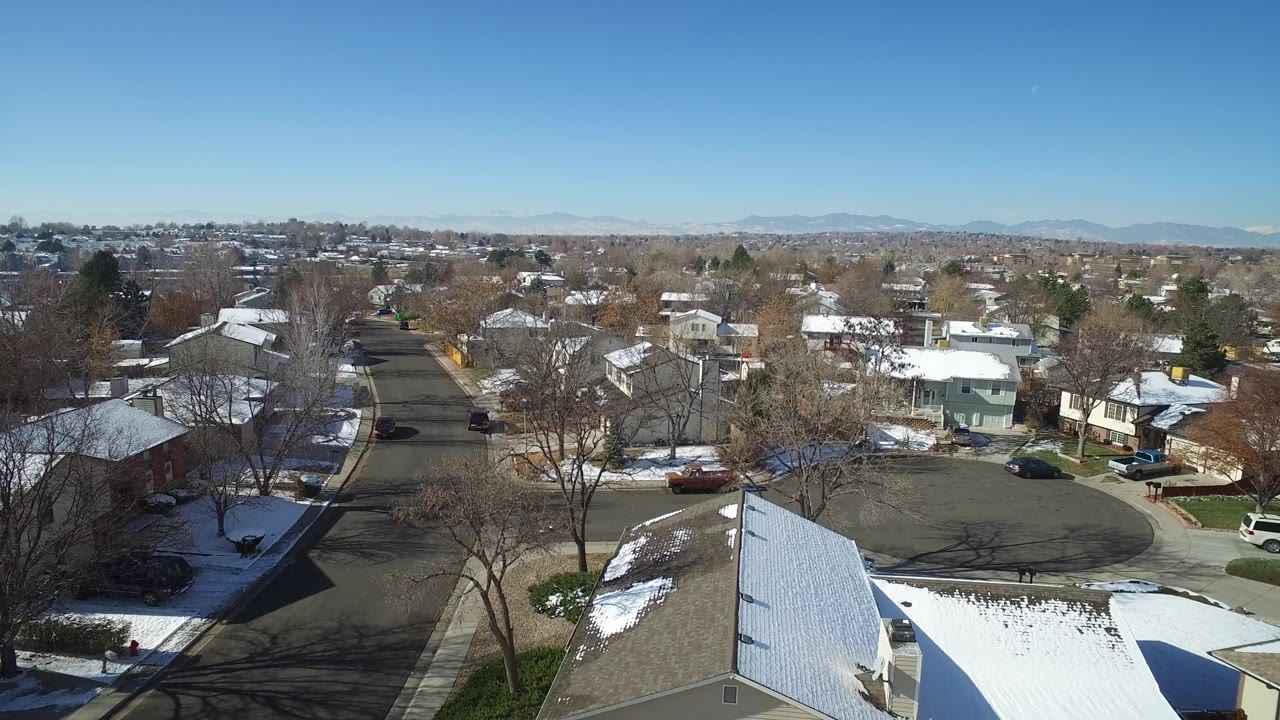 Wide aerial view of houses, bare trees, snow patches, and mountains.