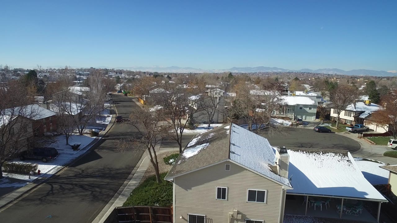 Elevated view over Hidden Lake, Colorado homes with snow on rooftops, bare trees, a residential street, distant mountains, and no visible people at the start of the clip.
