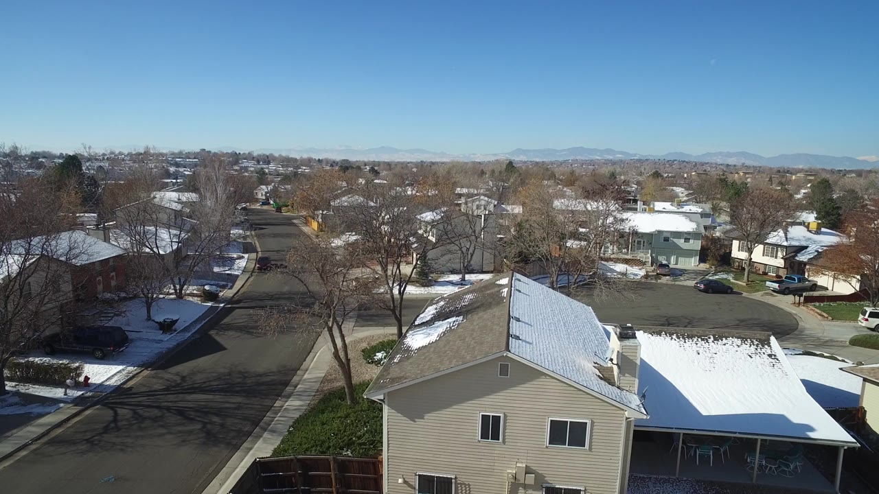 Drone-height frame in Hidden Lake, Colorado showing a beige house roof with snow, neighboring homes, leafless trees, parked cars, and no visible people.
