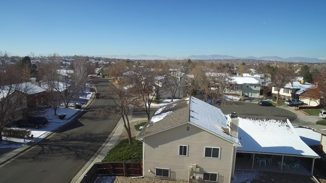 Snowy suburb with a beige house, carport roof, and distant hills.