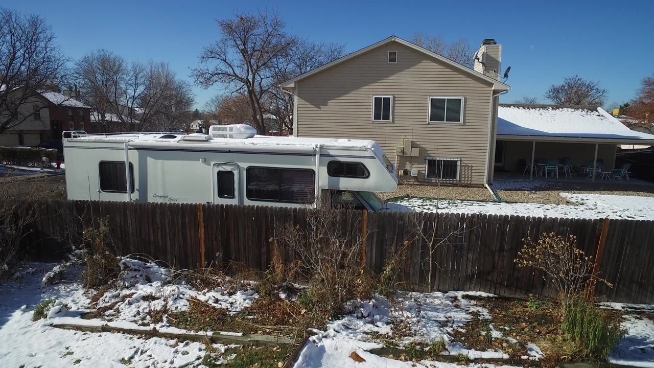 Snowy yard with wooden fence, white RV, and beige house behind it.