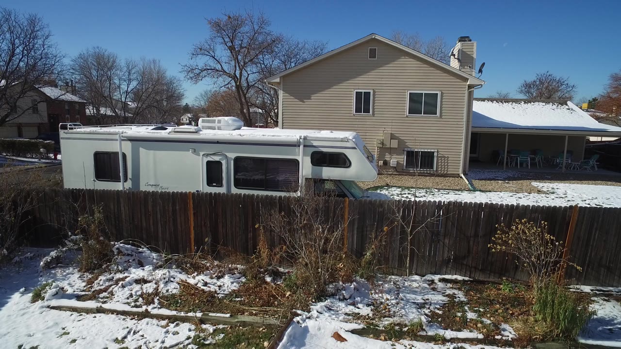 Hidden Lake, Colorado frame with the RV more centered behind the fence, the beige house roof and windows in back, snow patches across the lawn, and no visible people.