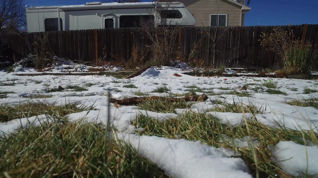Low winter yard frame in Hidden Lake, Colorado with uneven snow, exposed grass, a dark fence, a light RV or trailer, part of a house, and no visible people.