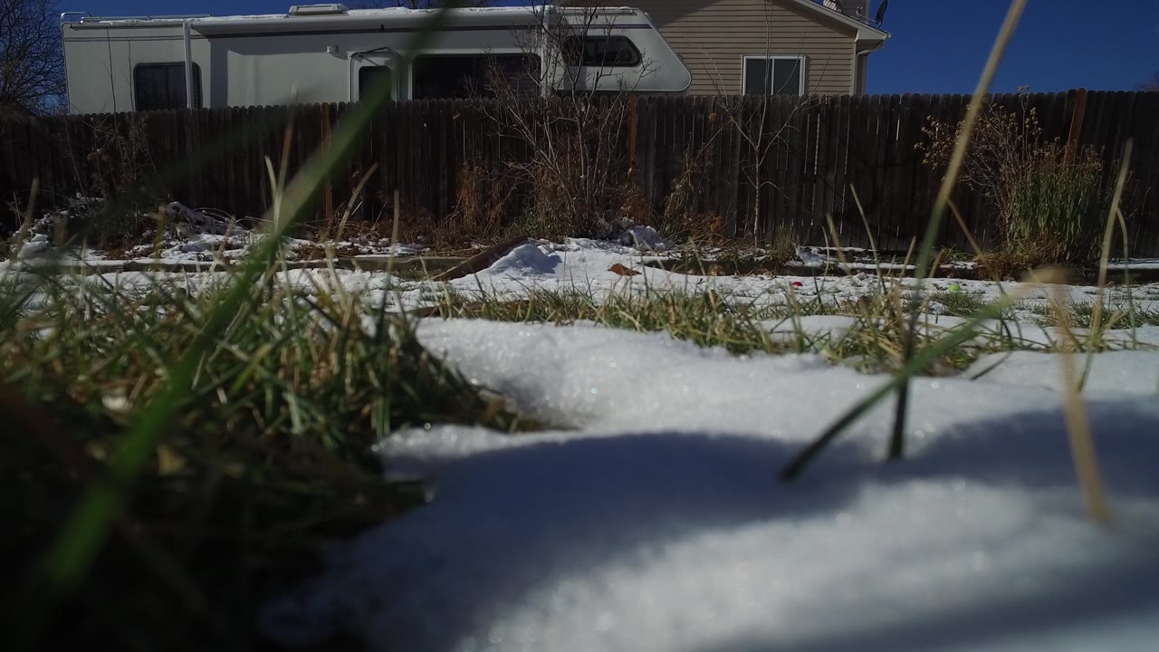 At 0:01 in Hidden Lake, Colorado, foreground blades of grass cross the lens above snow while a wood fence, white RV, and house remain in the background, with no visible people.