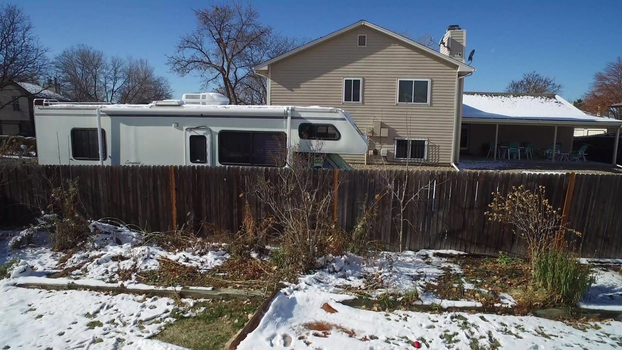 Aerial frame in Hidden Lake, Colorado showing a white RV parked behind a wood fence beside a beige two-story house, with snow patches, leafless trees, and no visible people.