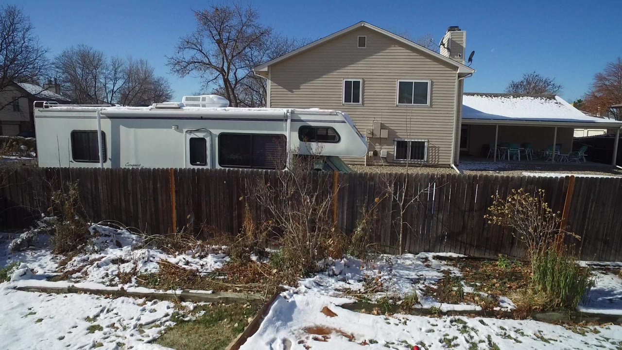 Aerial frame in Hidden Lake, Colorado with the white RV centered behind the fence, the beige house behind it, snow on the yard, bare trees, and no visible people.