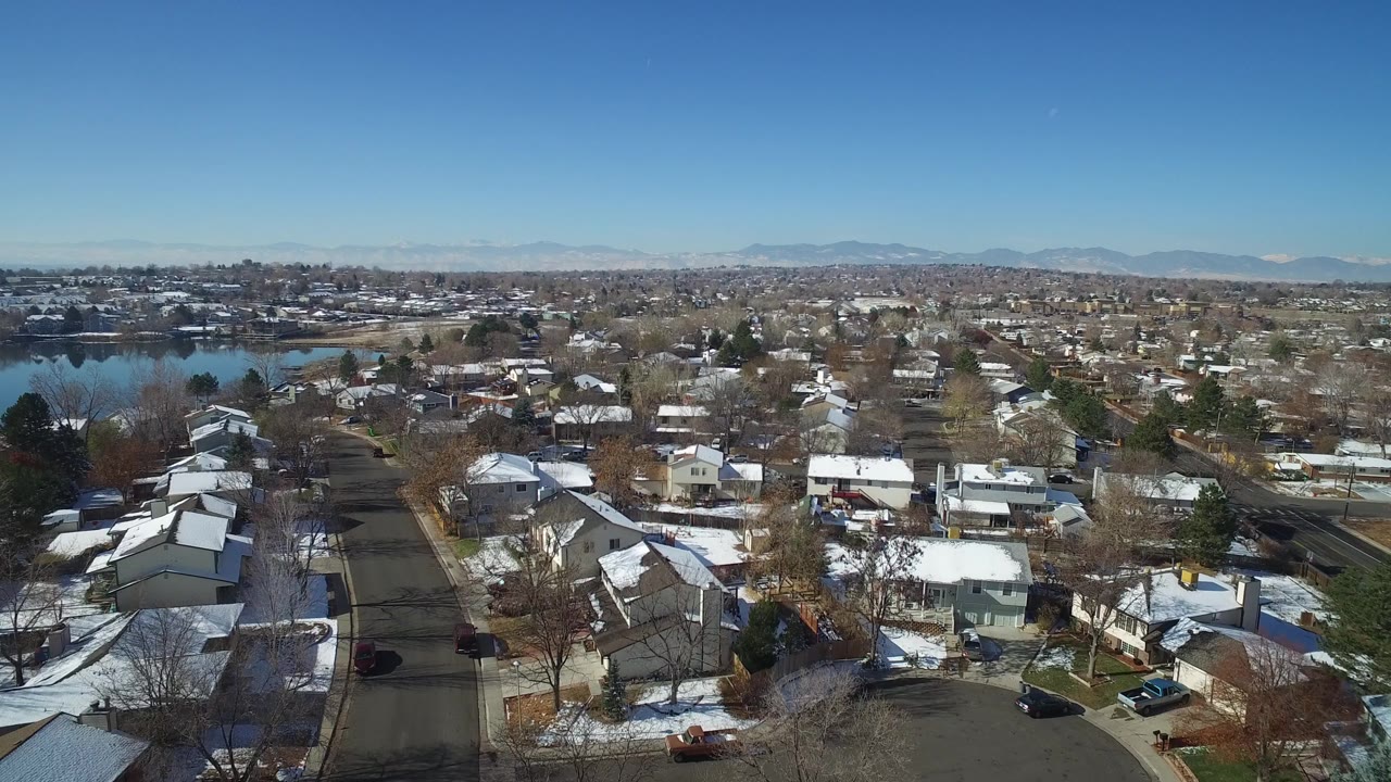 Second aerial frame in Hidden Lake, Colorado, with a curving residential street in the foreground, snow on roofs and yards, the lake left of frame, and no visible people.