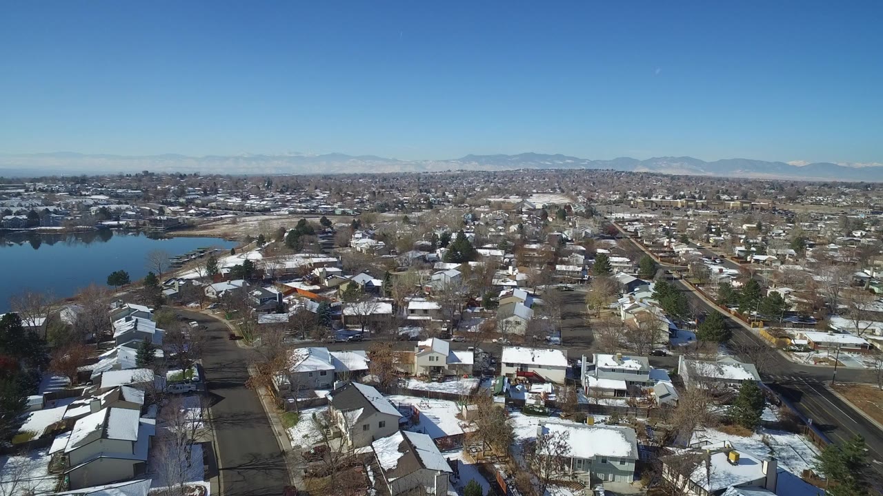 Aerial view of houses, snowy rooftops, streets, lake water, and distant mountains.