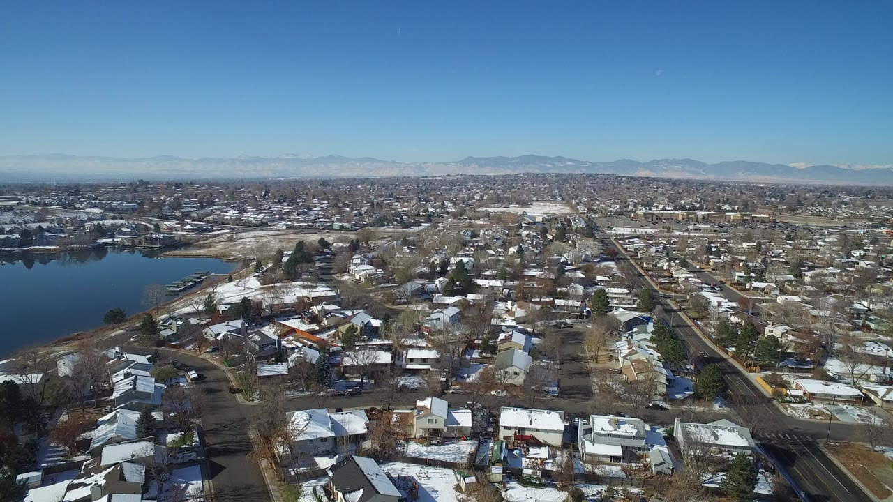 Aerial frame over Hidden Lake, Colorado, showing snow-dusted rooftops, a dark lake on the left, gridded streets, distant mountains, blue sky, and no visible people.