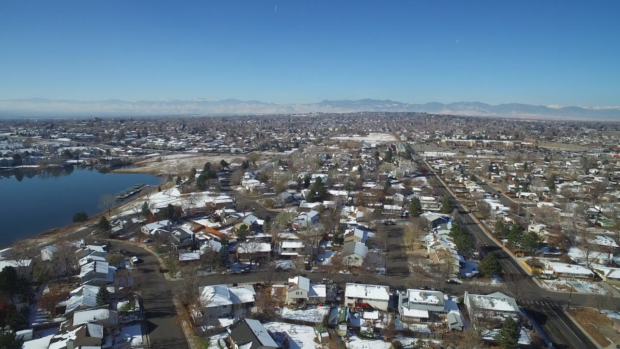 Aerial view over snow-dusted houses beside a lake and distant mountains.
