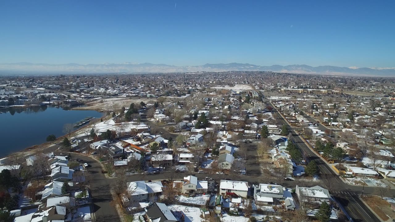 A second aerial moment over Hidden Lake, Colorado, shows clustered homes, gridded roads, bare trees, the lake along the left edge, distant sprawl, low mountains, and no visible people.