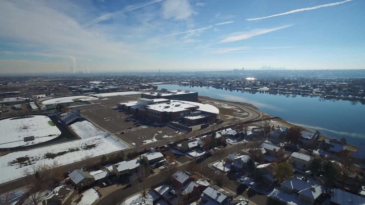 Aerial frame from Hidden Lake, Colorado showing snow-dusted houses in the foreground, large low buildings near the water, a blue lake, distant mountains, and no visible people.